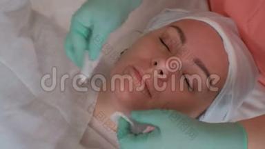 一个戴着蓝色手套的美容师用棉垫擦着一个年轻女人的脸。 美容界的美女
